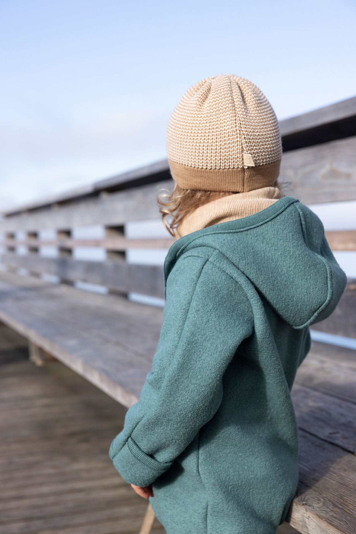 Child viewed from behind, wearing a blue boiled merino wool outfit and a beige beanie on a wooden deck