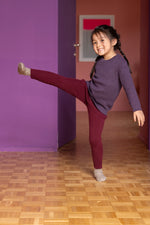 Young girl in a purple sweater and red Disana merino wool leggings posing in a room with purple walls and a wooden floor.