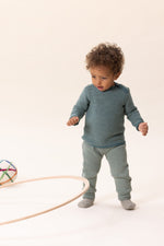 Child in teal merino wool sweater standing on a white background with toys around