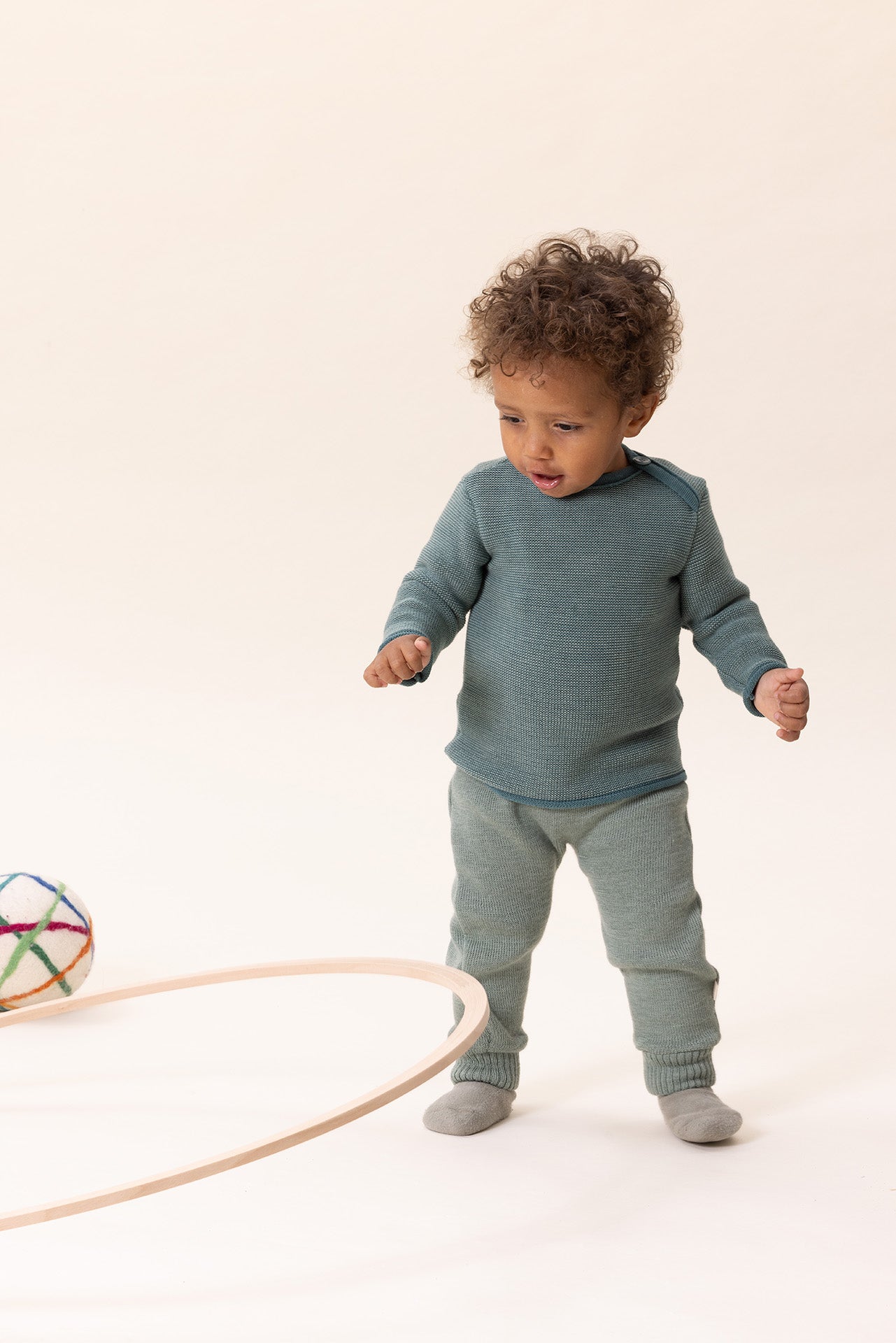 Child in teal merino wool sweater standing on a white background with toys around