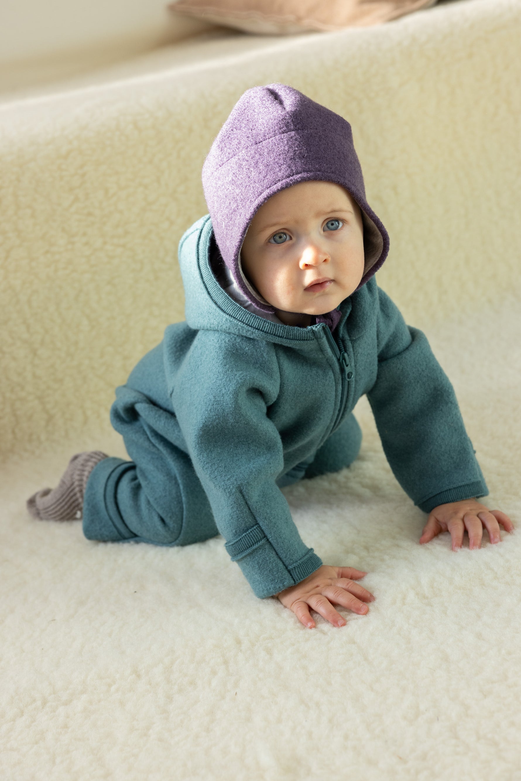 Baby in teal Disana boiled merino wool suit and purple hat on a beige surface