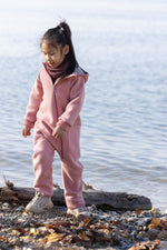 Child in a pink Disana boiled wool suit standing on a log by a body of water