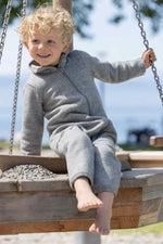 Child in a gray merino wool outdoor suit sitting on a wooden swing set with a blurred background