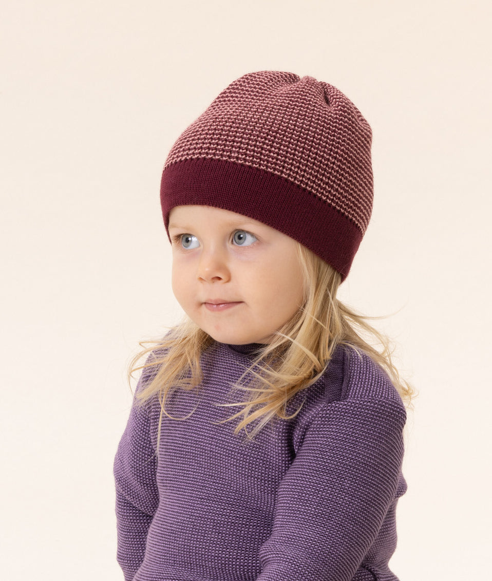 Child wearing a red knit merino hat and sweater from Disana on a beige studio background