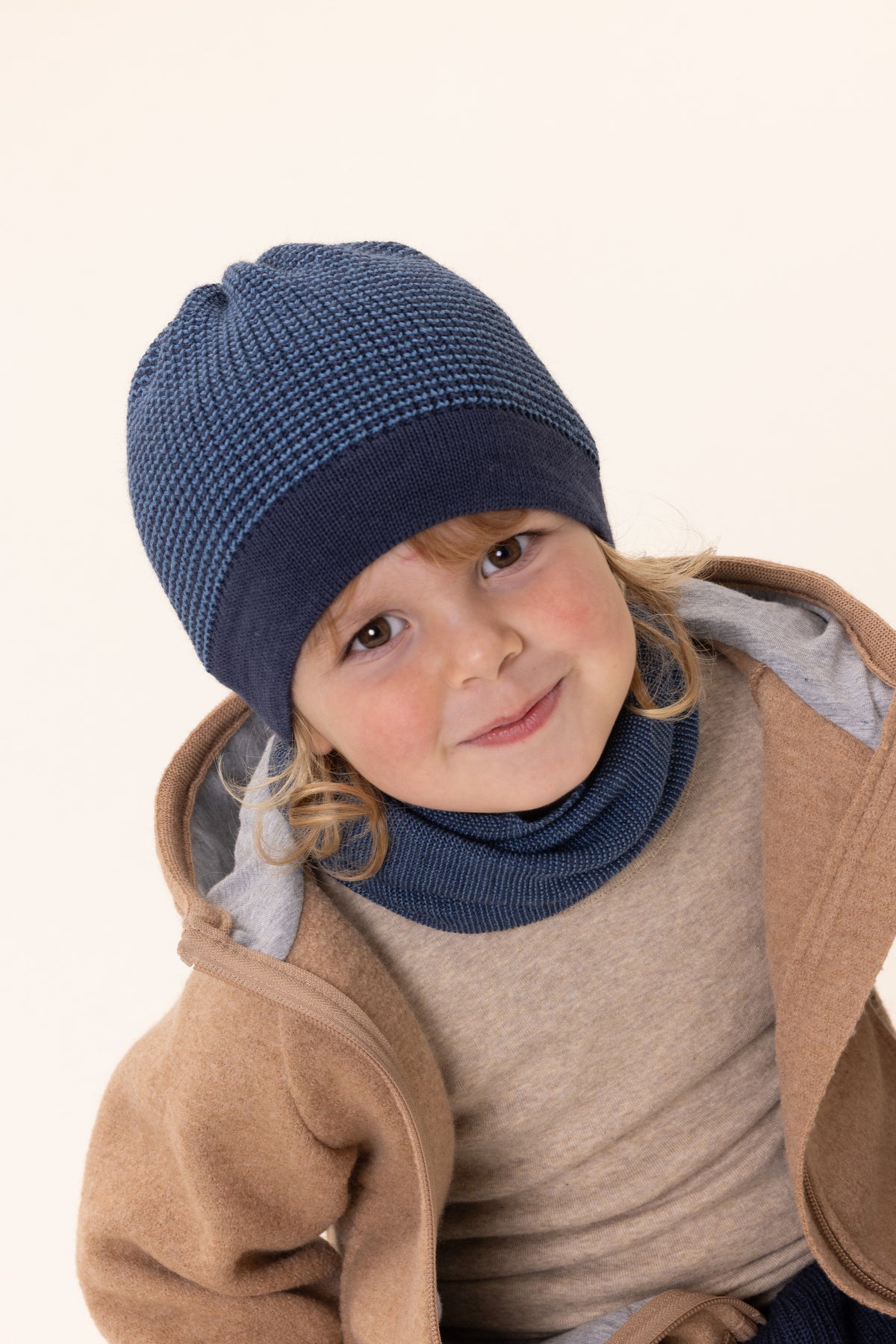 Child wearing a blue knit hat and merino wool scarf from Disana on a beige background