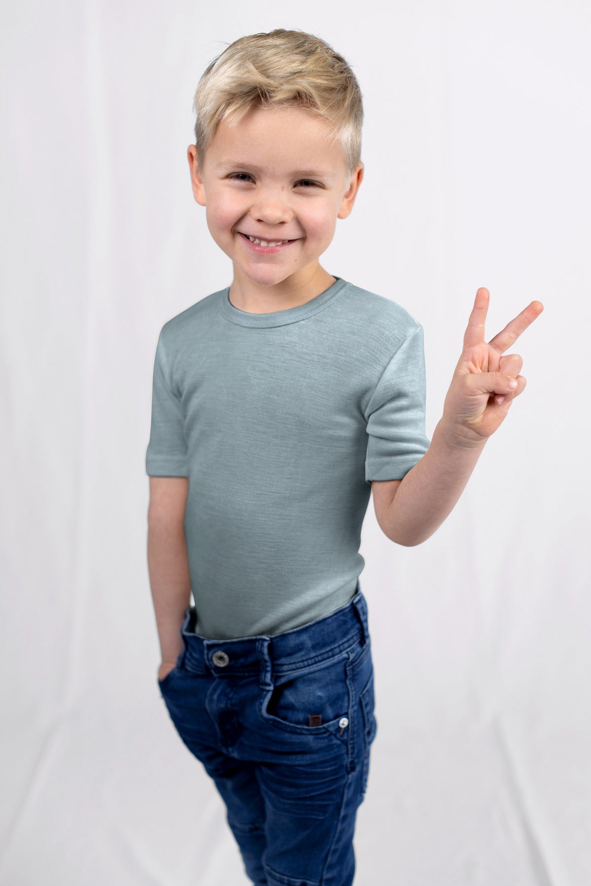 Child wearing a light blue merino wool shirt from Engel and dark blue jeans, making a peace sign on a white background