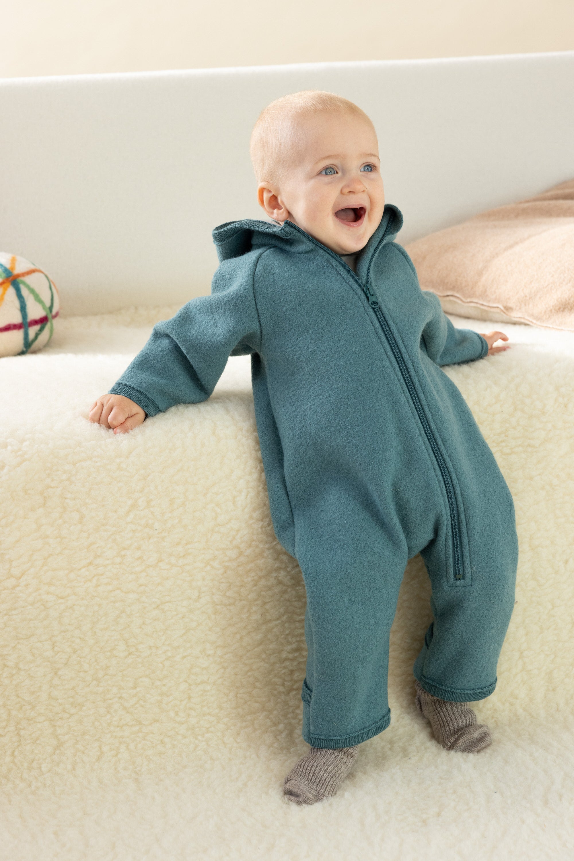Baby in a teal onesie sitting on a white surface with a colorful ball in the background
