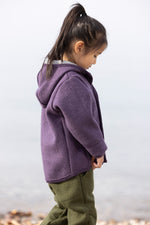 Child wearing a purple hoodie and green pants standing on a beach with a white background