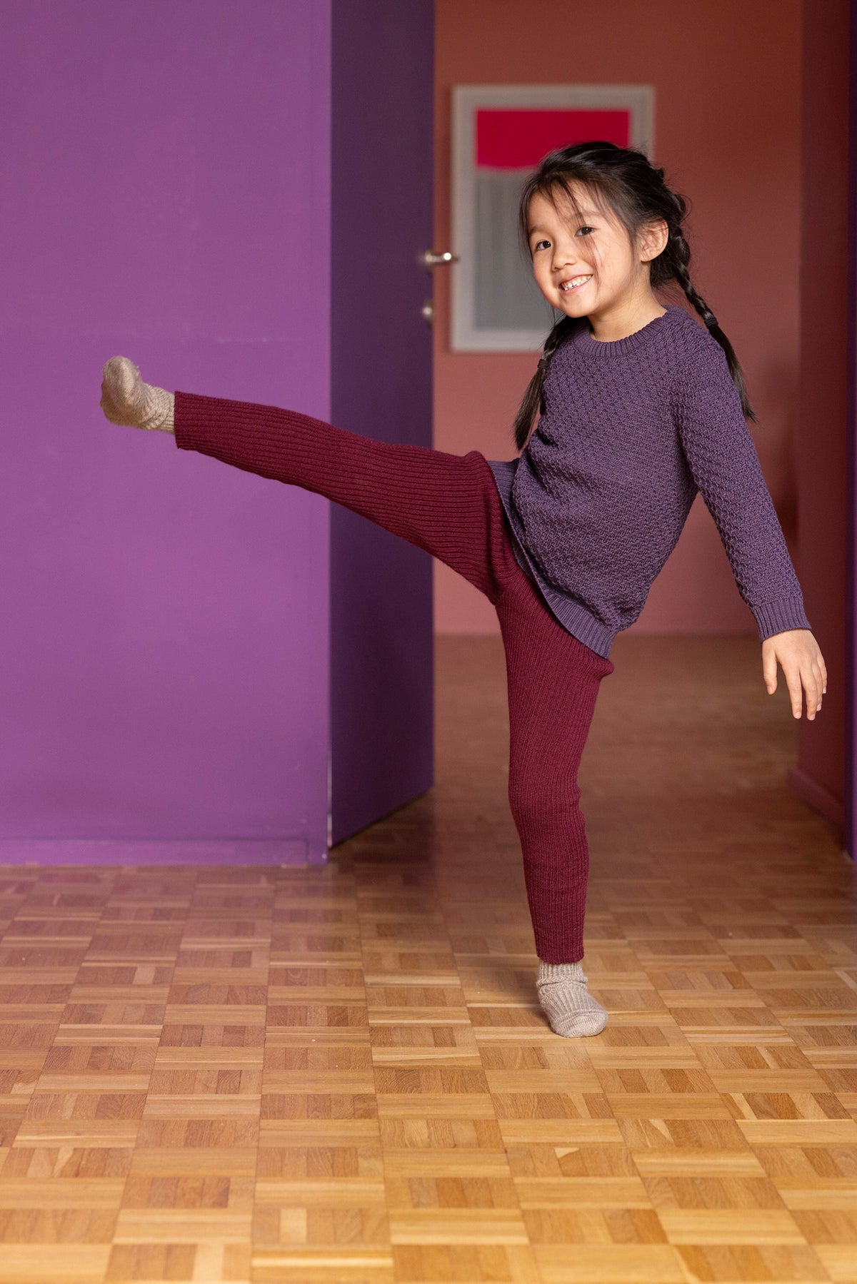 Young girl in a purple sweater and red pants posing in a room with purple walls and a wooden floor.