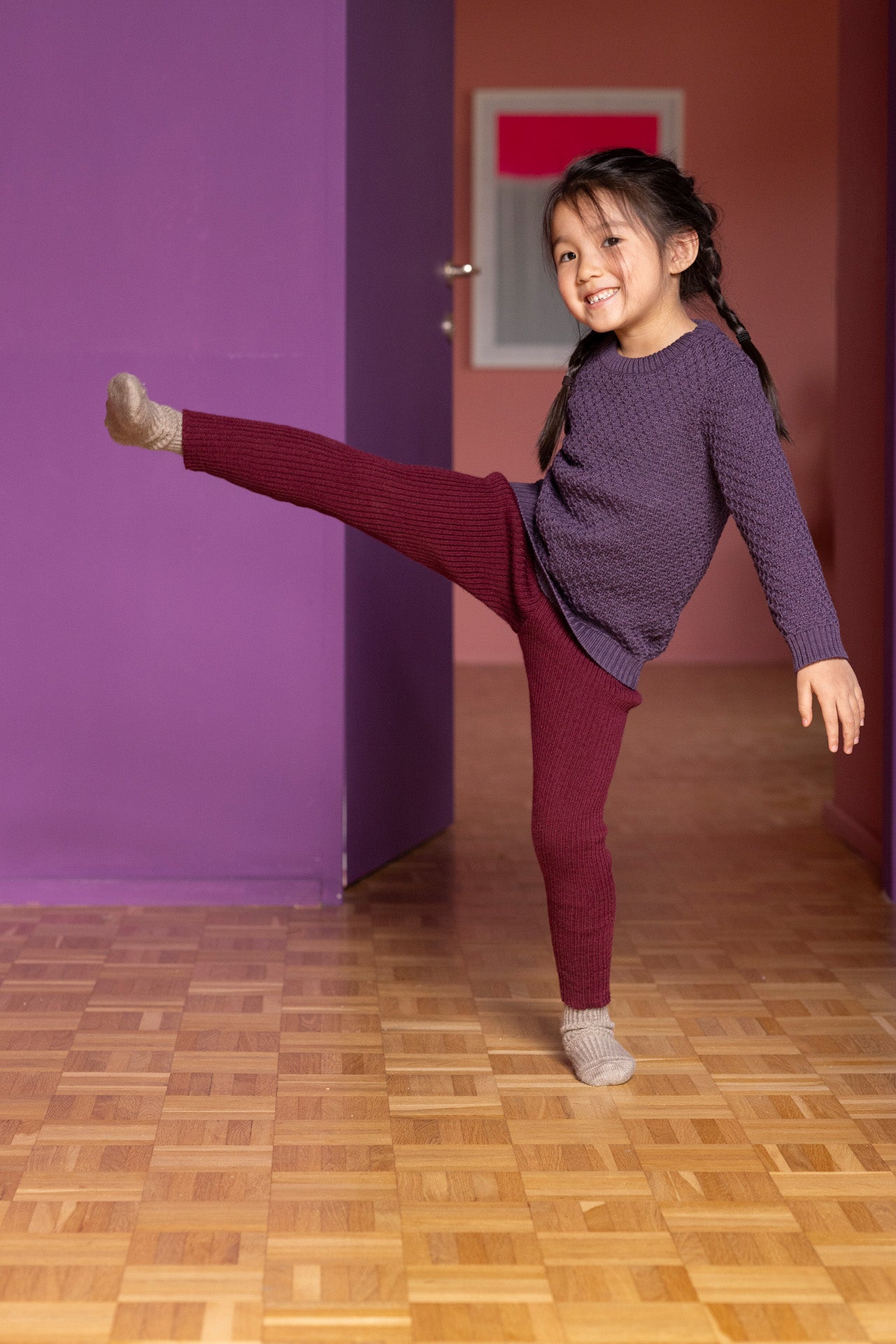Young girl in a purple sweater and red pants posing in a room with purple walls and a wooden floor.