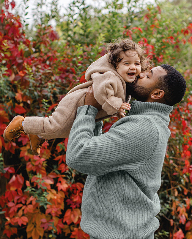 A man in a ribbed sage green turtleneck sweater holds and kisses a young child outdoors in front of a dense bush with vibrant red and orange autumn leaves.