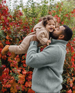 A man in a ribbed sage green turtleneck sweater holds and kisses a young child outdoors in front of a dense bush with vibrant red and orange autumn leaves.