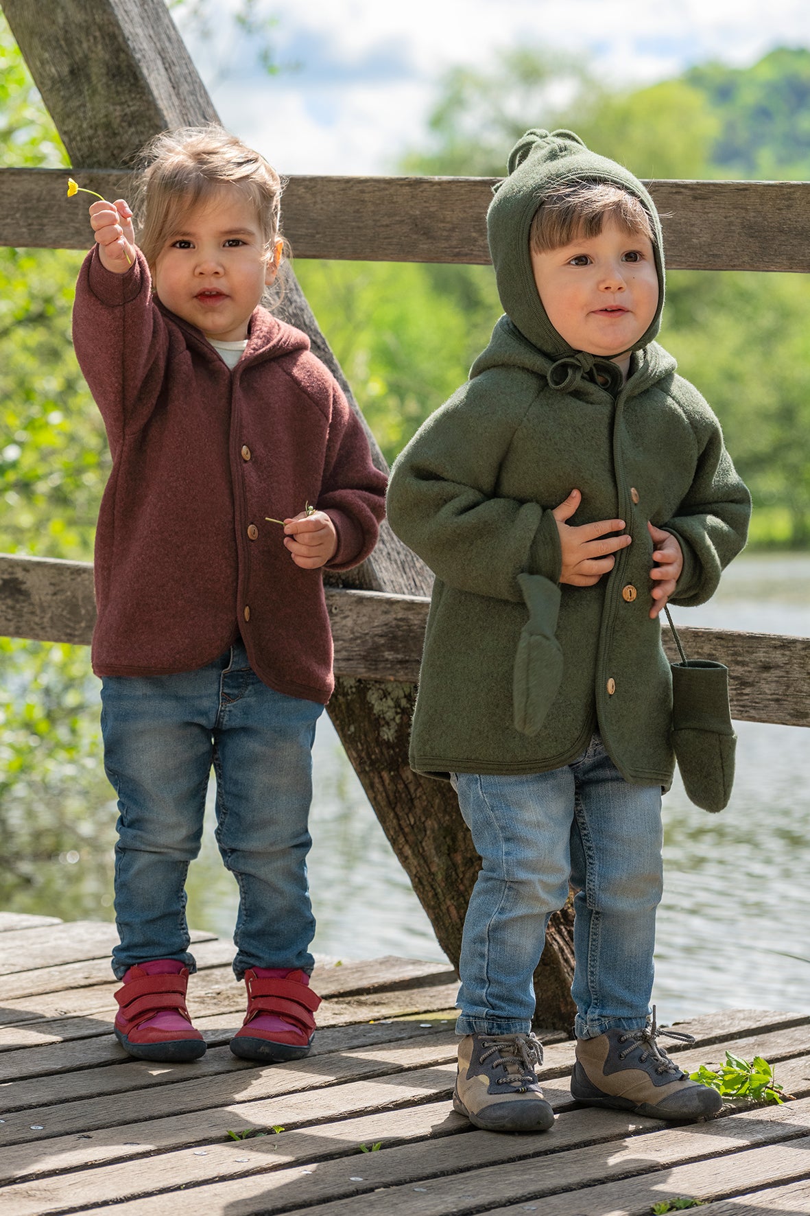 Two young children standing on a wooden dock outdoors by the water. They are wearing wool fleece jackets in brown and green.